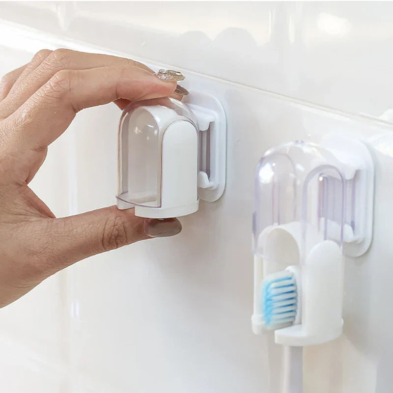 Hand holds a detached wall-mounted toothbrush holder near its adhesive mount; an identical holder holding a white-handled blue-bristled toothbrush sits to its right.