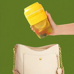 Electric Citrus Juicer - A hand holds a tilted yellow cup with fruit and drink inside, above a white shoulder bag against green background