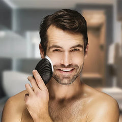 Shirtless man using a black ribbed facial cleansing brush on his cheek, in a bathroom setting