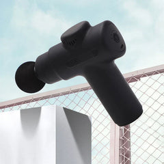 Electric Fascia Vibration Recovery Gun - Black mini massage gun resting on a white block at an angle against a chain-link fence and sky, with a round massage head attached