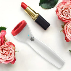 Electric Lip Plumper Enhancer Device - Lip plumping tool laid flat on white surface next to a red lipstick, surrounded by pink rose blooms and green leaves