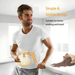 A man in a white shirt holds a glass self-stirring mug with a wooden lid and handle in a bright kitchen, with spilled coffee beans on a counter nearby.
