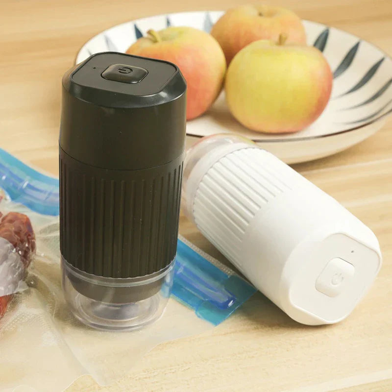 Black and white handheld electric pumps rest on a table beside apples, with vacuum storage bags with electric pump partially visible nearby