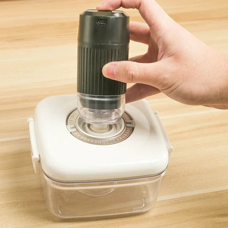 Hand holding a small black electric pump over the valve of a square vacuum storage container on a wooden surface.