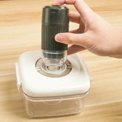 Hand holding a small black electric pump over the valve of a square vacuum storage container on a wooden surface.
