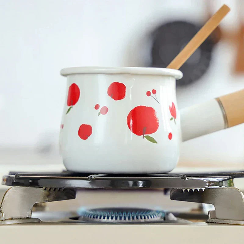 White enamel pot with red apple designs sits on a gas stove burner; a heat diffuser plate is not visibly present.