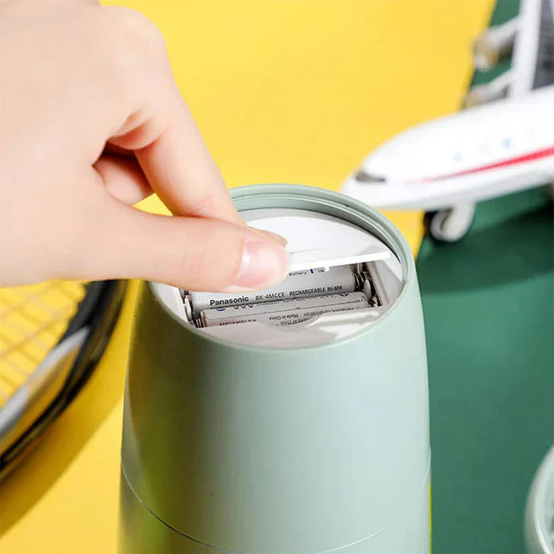 Suction Tumbler Mug - A finger lifts the battery compartment cover on a light green cylindrical device, revealing two installed AA rechargeable batteries.