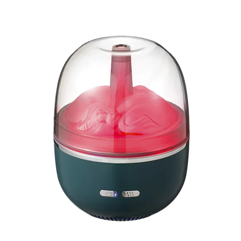 Un difusor de aceites esenciales humidificador con cúpula transparente y base verde azulado, con luz interior rosa, boquilla de vapor y dos botones de control en su base.