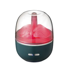 Un difusor de aceites esenciales humidificador con cúpula transparente y base verde azulado, con luz interior rosa, boquilla de vapor y dos botones de control en su base.