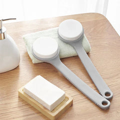 Loofah - Two long-handled bath brushes resting on a folded light green washcloth, arranged on a wooden countertop alongside bar soap and a soap dispenser