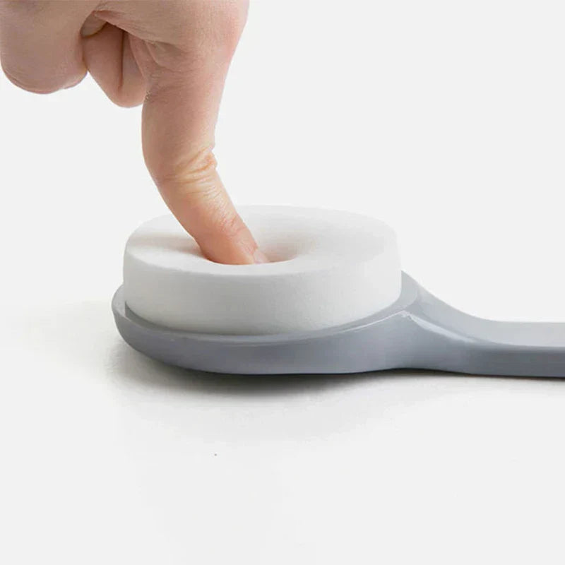 Loofah - A finger presses into the soft white sponge head of the gray-handled bath brush