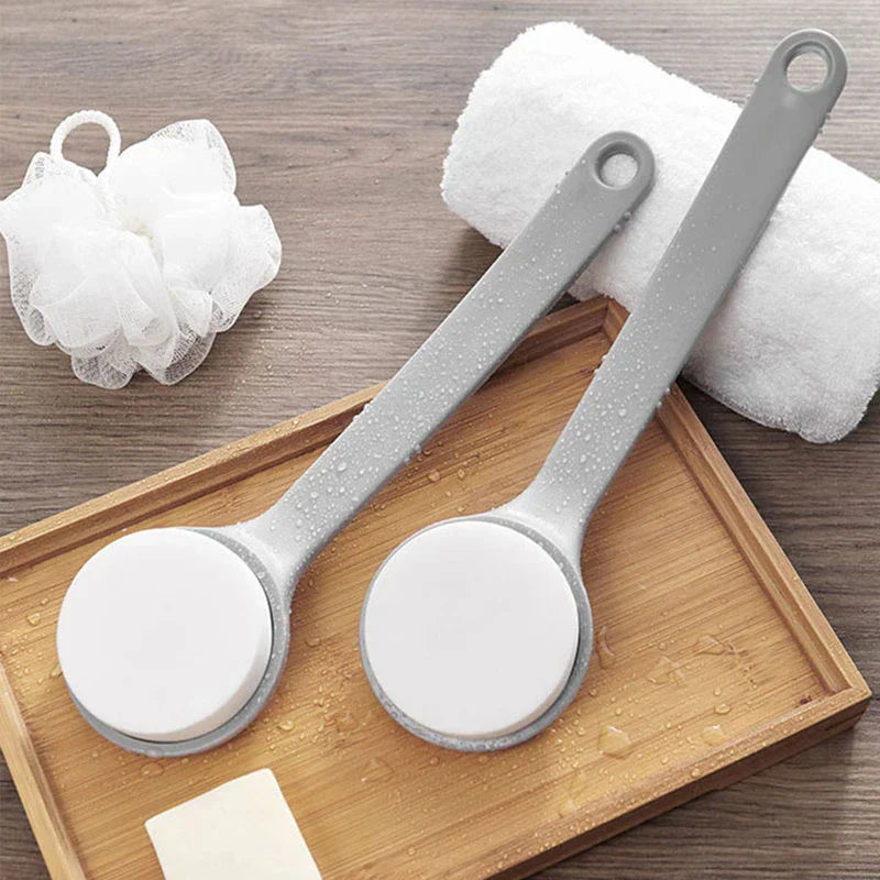 Loofah - Two long-handled bath brushes with water droplets rest on a wooden tray, alongside a rolled towel, bath pouf, and small soap piece