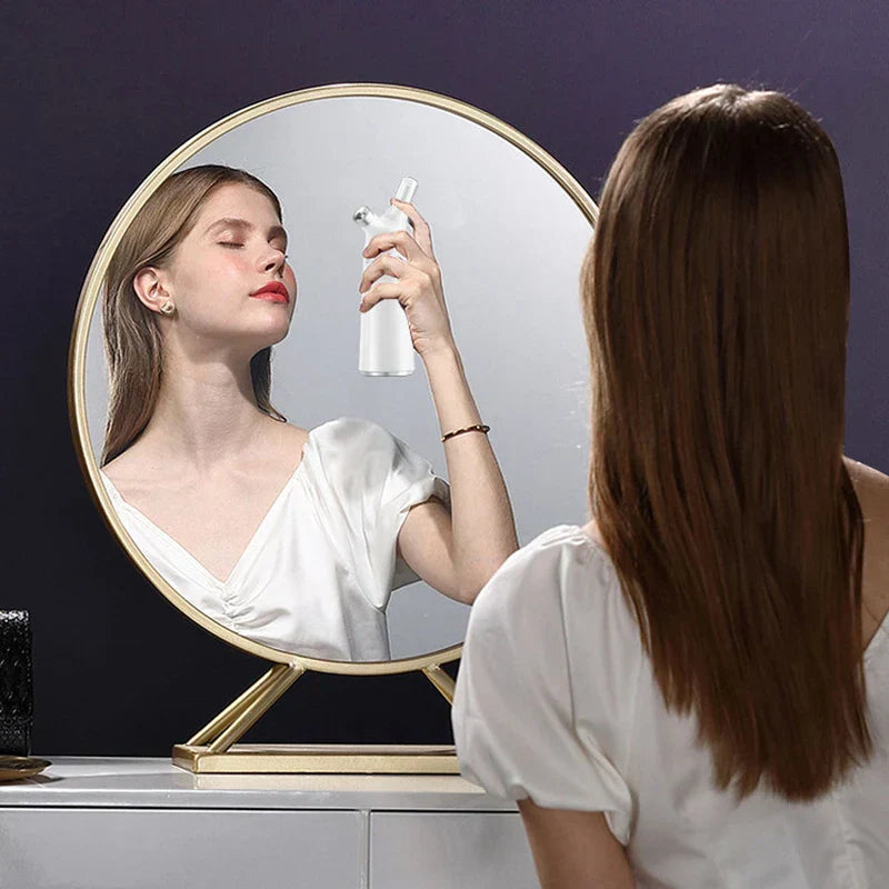 Nano Mist Sprayer - Woman facing a round gold-framed mirror, holding the white spray device to her face while viewed from behind.
