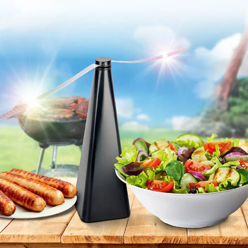 fliegenvertreiber ventilator - Schwarzes konisches Gerät auf einem hölzernen Picknicktisch neben gegrillten Würstchen und einer frischen Salatschüssel, mit einem Holzkohlegrill im sonnigen Außenbereich im Hintergrund