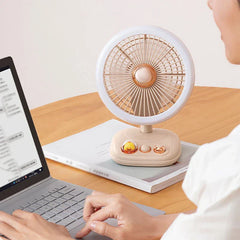A person types at a laptop while a portable standing fan rests on a wooden desk in front of them.