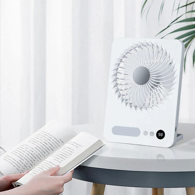 Tischventilator - Weißer Tischventilator steht auf einem runden Tisch neben einem aufgeschlagenen Buch, das von Händen gehalten wird, weißer Vorhang und Pflanze im Hintergrund