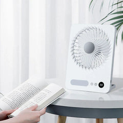 Tischventilator - Weißer Tischventilator steht auf einem runden Tisch neben einem aufgeschlagenen Buch, das von Händen gehalten wird, weißer Vorhang und Pflanze im Hintergrund