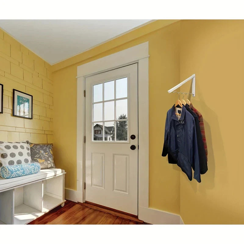 perchero retráctil para secar ropa - Perchero de pared plegable en color blanco montado en una pared amarilla cerca de una puerta, sosteniendo tres chaquetas