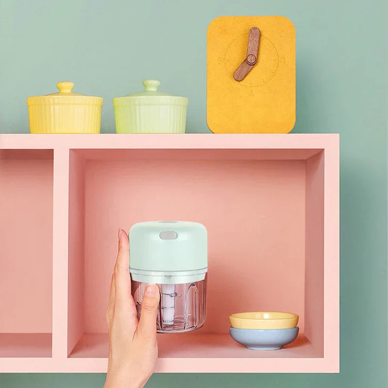 Light green mini electric chopper is held by a hand, placed inside a pink shelf cubby with small bowls and a clock on the shelf above