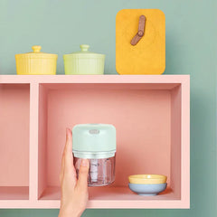 Light green mini electric chopper is held by a hand, placed inside a pink shelf cubby with small bowls and a clock on the shelf above