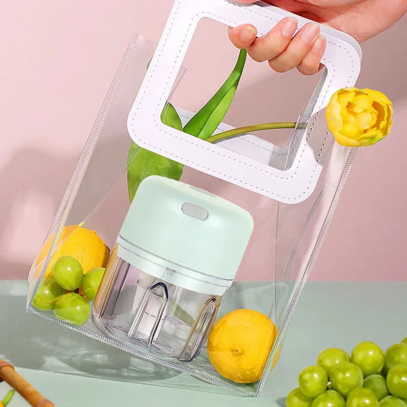 A mint green electric mini chopper sits inside a clear tote bag with lemons, green grapes, and a yellow tulip beside extra fruit on a pale table.
