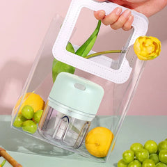A mint green electric mini chopper sits inside a clear tote bag with lemons, green grapes, and a yellow tulip beside extra fruit on a pale table.