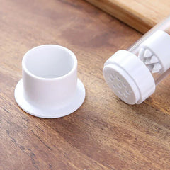 Garlic Press - Two white plastic parts on a wooden surface: a short cylindrical cap, and a perforated component with internal spikes attached to a clear tube.