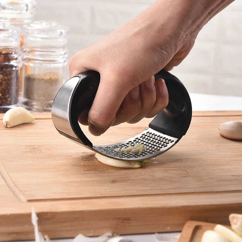 Garlic Mincer Tool - Hand holding a curved garlic press, pressing a garlic clove on a wooden cutting board with garlic pieces visible