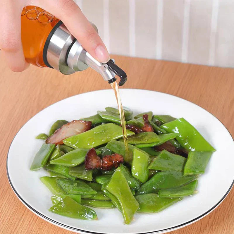 Glass Cruet - Hand holding an oil bottle pouring amber oil onto a plate of cooked green vegetables and sliced meat, on a wooden table.