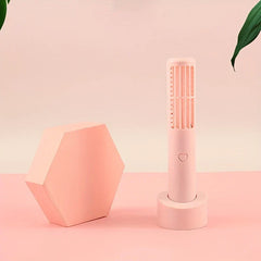 Mini Fan - Light pink handheld tower fan on its stand, beside a leaning hexagonal block, with partial green leaves at top corners