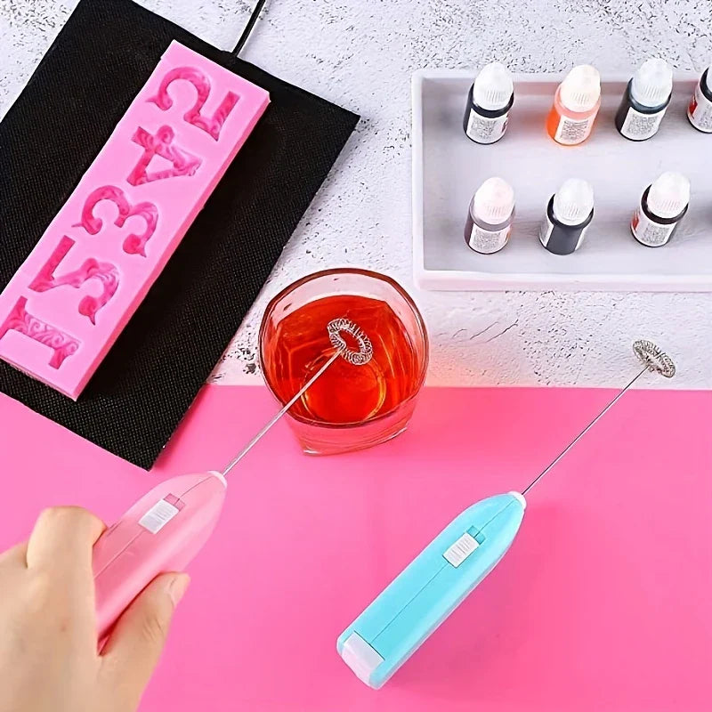 Handheld Electric Milk Frother - A hand uses a pink electric stirrer in a glass of red liquid, with a blue stirrer, number mold and dye bottles on a tray nearby