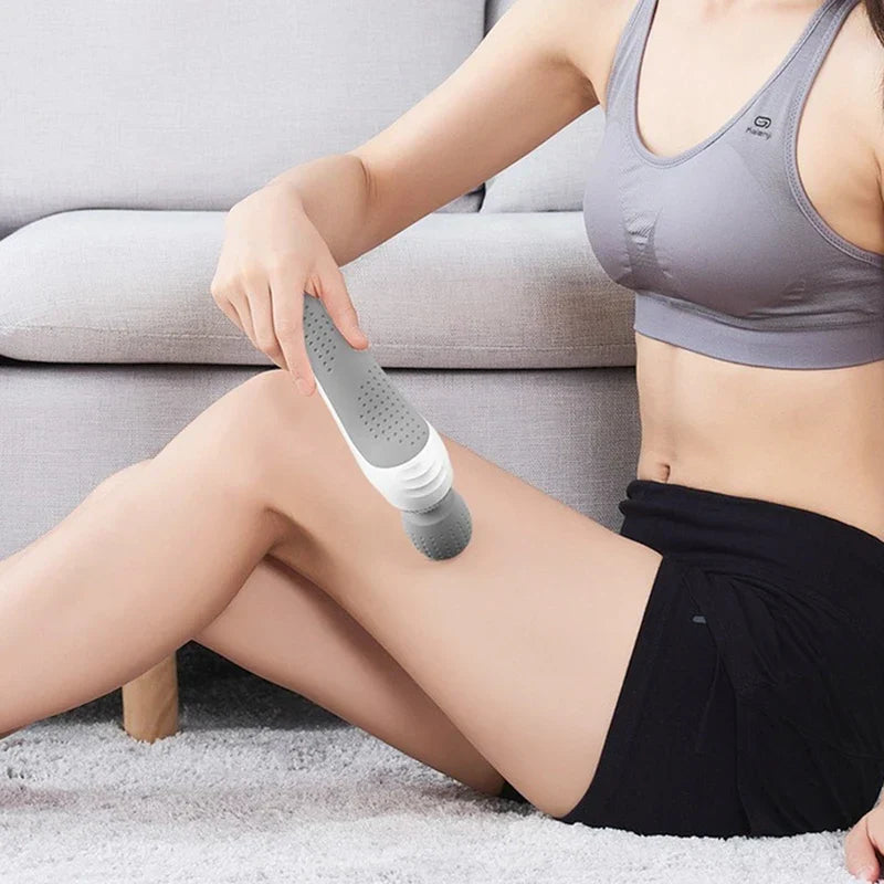 handmassagegerät für den körper - Person in Sportkleidung hält ein grau-weißes Massagegerät gegen den Oberschenkel, sitzend auf Teppich neben einem Sofa
