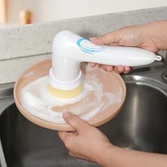 Electric Spin Scrubber - Person using the handheld cleaning tool with a sponge attachment to scrub a soapy peach-colored plate over a stainless steel sink