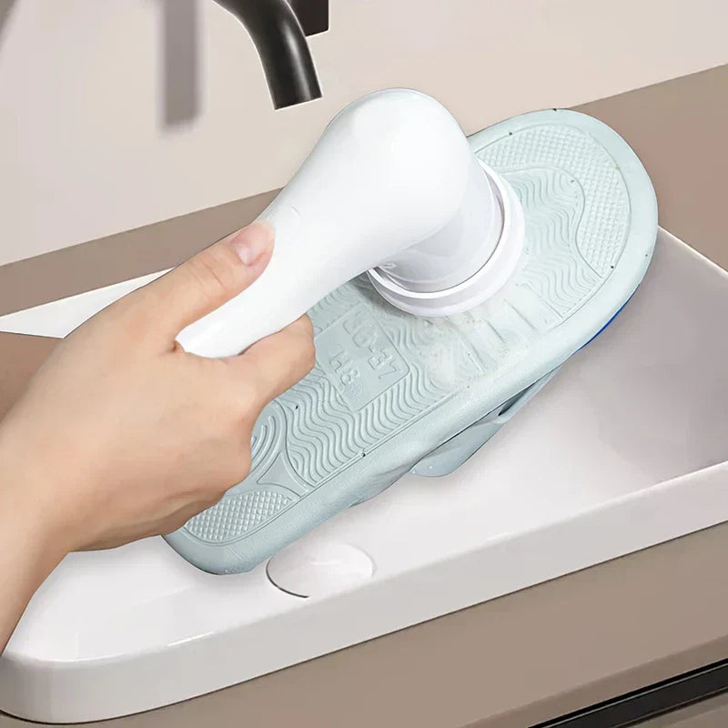 Electric Spin Scrubber - A hand holds a white-handled cleaning brush scrubbing a light blue sandal sole in a white sink, with a dark faucet above.