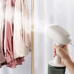 vaporizador de prendas - Mano sosteniendo un vaporizador blanco que emite vapor hacia camisas colgadas