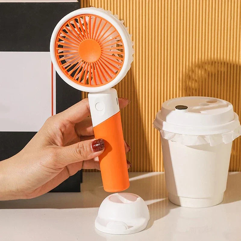 Mini Fan - Hand holding an orange and white handheld fan, detached base and cup on the table