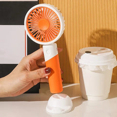 Mini Fan - Hand holding an orange and white handheld fan, detached base and cup on the table