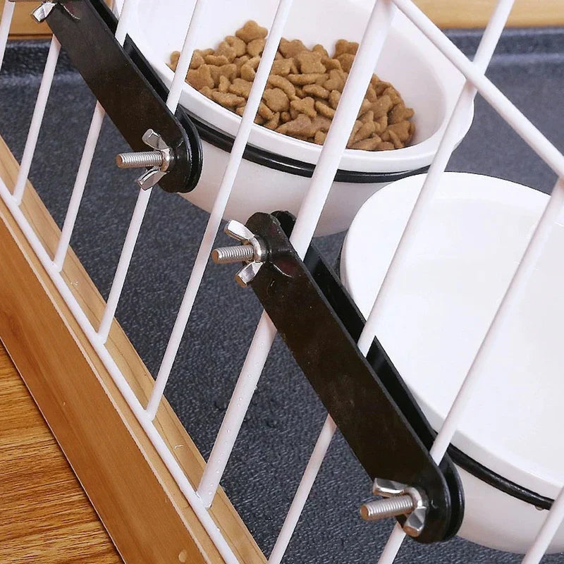 Two white bowls secured to a white wire pet crate, one with brown kibble, and a pet crate water bowl is mounted at the lower position.