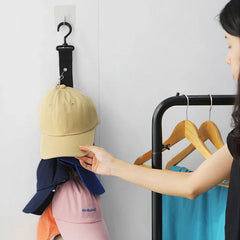 Hat Organizer - Woman's hand reaching for a beige baseball cap hung on a black strap hanger mounted to a white wall, with a clothing rack of wooden hangers beside her
