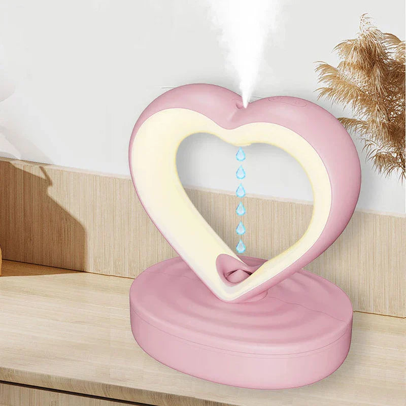 Humidifier - Pink heart-shaped device in use, emitting mist from its top with water droplet graphics inside its open center, placed on a wood surface.