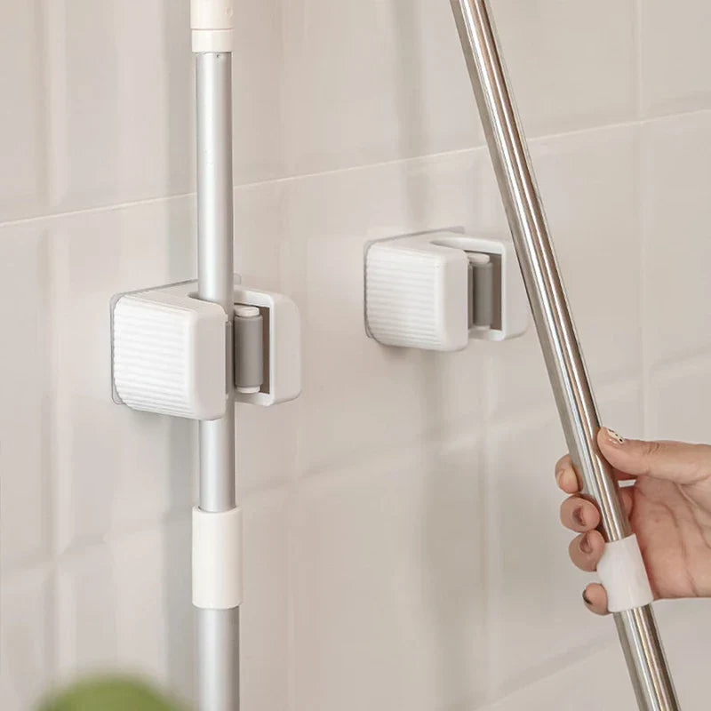 Mop Broom Holder - Two white wall hooks mounted on light tiled wall, one holding a silver cleaning pole, a hand positioning a second silver pole near the empty hook