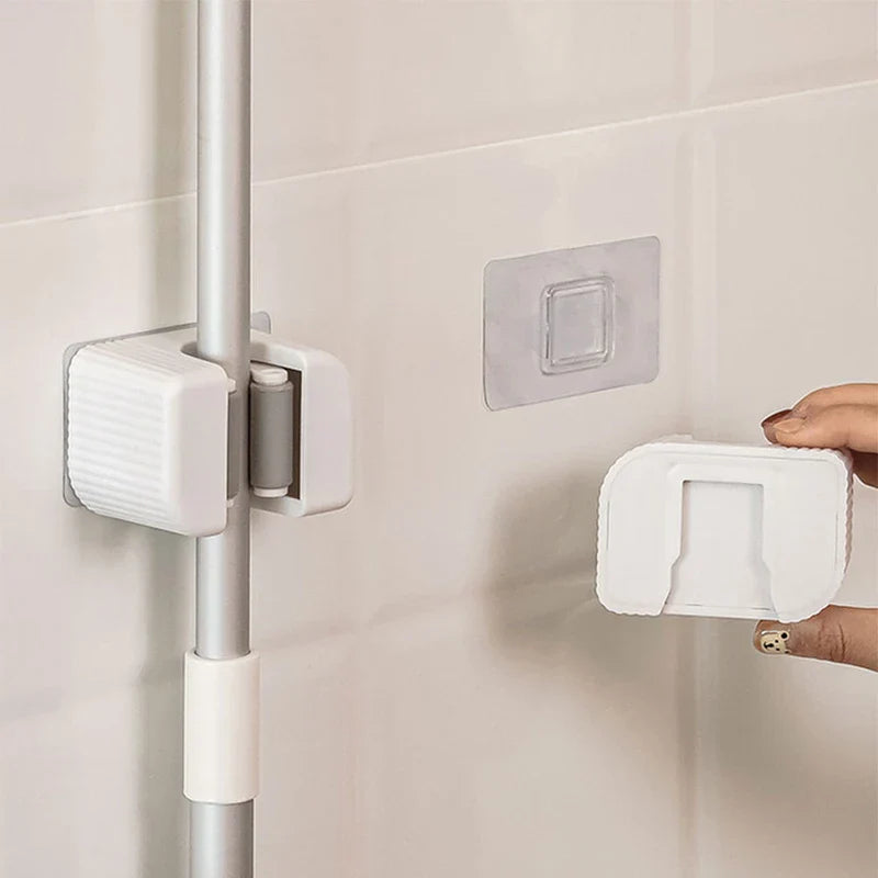 Soporte para trapeador y escoba: un gancho de pared blanco sosteniendo un palo de limpieza plateado en una pared de azulejos, una mano sosteniendo un segundo gancho junto a su almohadilla adhesiva transparente de montaje