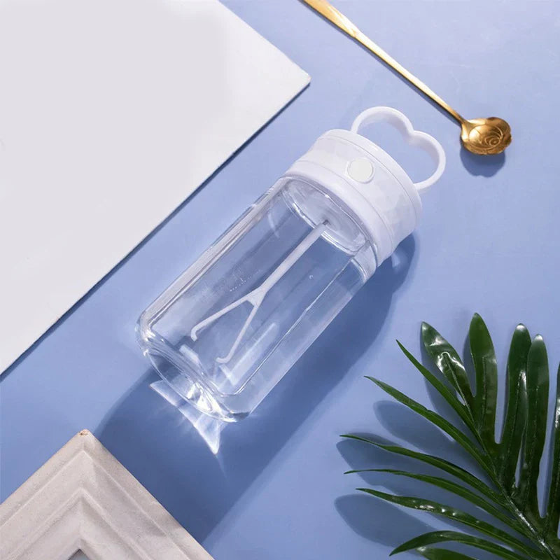 taza de viaje aislada para café - Botella de agua de plástico transparente acostada de lado con tapa blanca y asa en forma de corazón, junto a una cuchara dorada, bloc de notas blanco y hojas verdes sobre superficie azul claro
