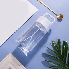 taza de viaje aislada para café - Botella de agua de plástico transparente acostada de lado con tapa blanca y asa en forma de corazón, junto a una cuchara dorada, bloc de notas blanco y hojas verdes sobre superficie azul claro