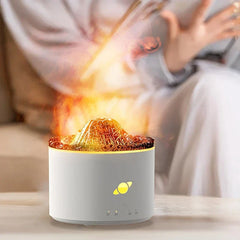 difusor humidificador de llama - Humidificador cilíndrico blanco con parte superior en forma de volcán iluminada que emite niebla, colocado sobre una mesa clara con una persona al fondo desenfocada