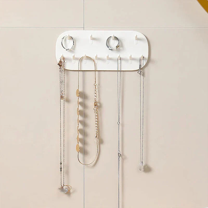 A wall-mounted jewelry storage organizer box with rings and several necklaces hanging from its hooks on a light tiled wall