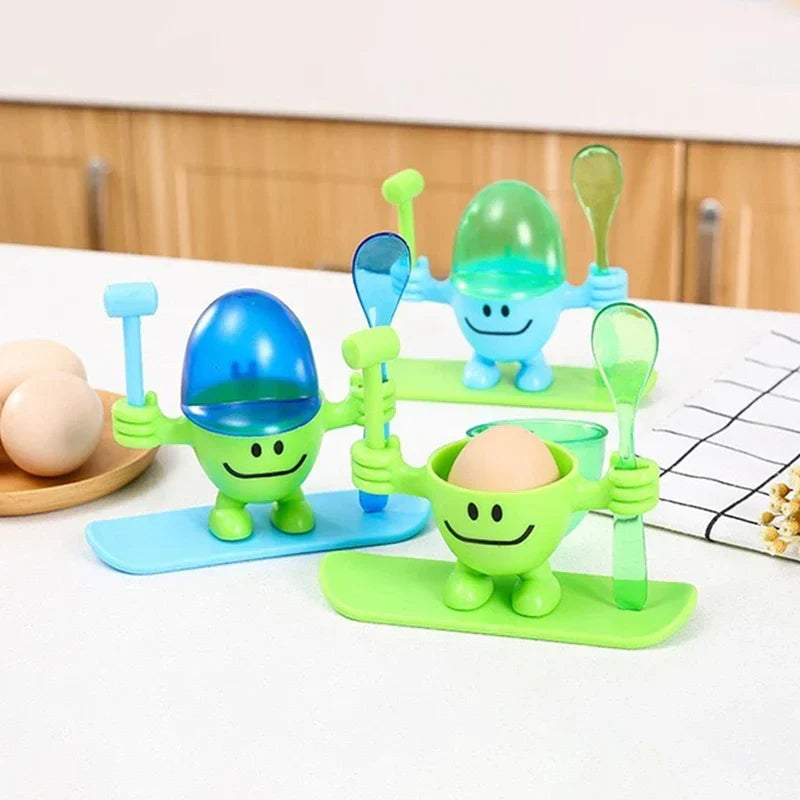 Egg Cup Holder - Three colorful novelty egg cup figurines with hammers and spoons, two with egg-shaped lids, one holding a brown egg, arranged on a countertop with whole eggs nearby.
