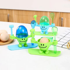 Egg Cup Holder - Three colorful novelty egg cup figurines with hammers and spoons, two with egg-shaped lids, one holding a brown egg, arranged on a countertop with whole eggs nearby.