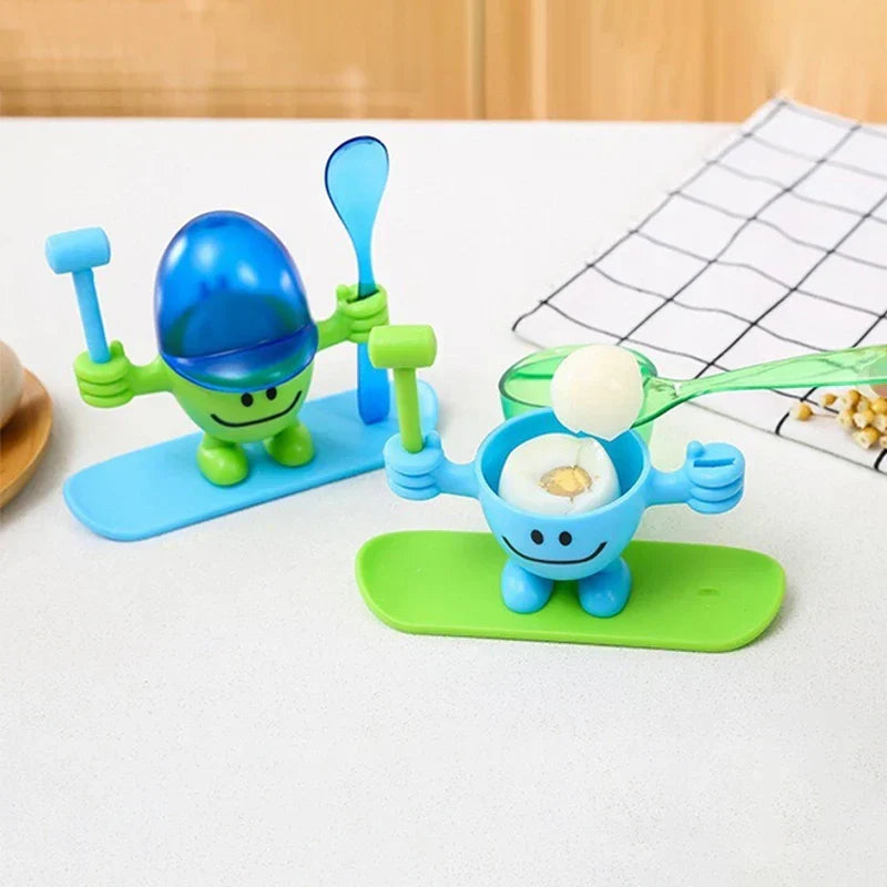 Egg Cup Holder - Two novelty egg cup figurines on colored bases; the blue cup holds a cracked soft-boiled egg, a spoon lifts a piece of egg white, the green cup has a blue lid.