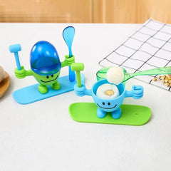 Egg Cup Holder - Two novelty egg cup figurines on colored bases; the blue cup holds a cracked soft-boiled egg, a spoon lifts a piece of egg white, the green cup has a blue lid.
